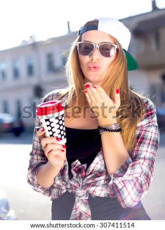 Summer outdoor fashion portrait of young stylish hipster cute girl drinking tasty coffee on the street and sending kisses ,casual trendy outfit, swag cap,checkered shirt and stylish sunglasses.