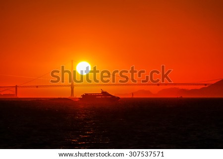 A sailboat and a ferryboat crossing the San Francisco Bay at sunset with the Golden Gate Bridge in the background.