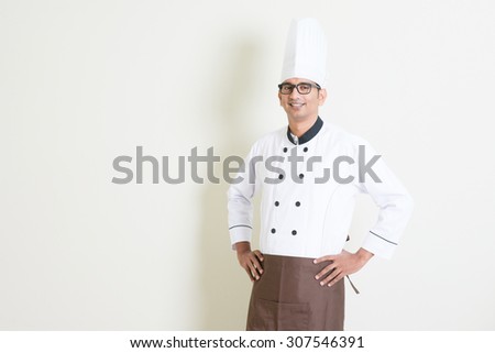Portrait of handsome Indian male chef in uniform hands on waist and smiling, standing on plain background with shadow, copy space on side.