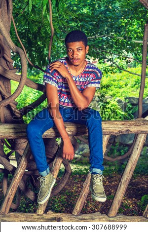 African American Man Relaxing on Park in New York. Wearing a colorful pattern shirt, blue jeans, sneakers, necklace, a young handsome guy is sitting on tree trunk fences, thinking. Instagram effect.
