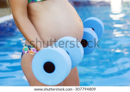 Beautiful pregnant woman is water aerobics with blue lightweight dumbbells near swimming pool 