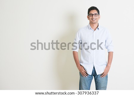 Portrait of handsome casual business Indian guy smiling, hands in pocket, standing on plain background with shadow, copy space at side.