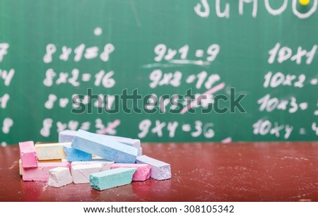 Blur texture background of school blackboard with written multiplication table and heap of colorful chalk blue pink violet, white yellow green colors lying on brown desk, horizontal picture