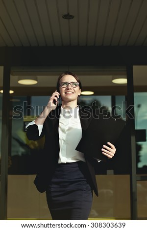 Happy smilling business woman coming out of office and talking on smartphone with documents in her hand. Successful business lady and professional lady walk around office.
