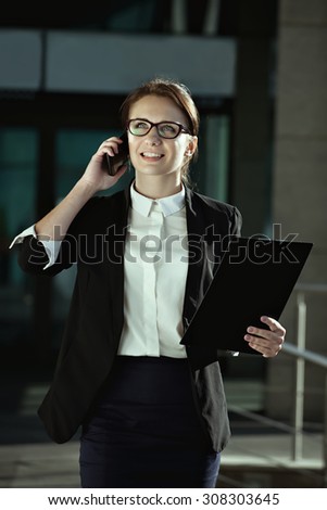 Business peolple concept- successful business woman talking on smartphone. Hppy young business lady manager with documents talking on phone on office industrial background.