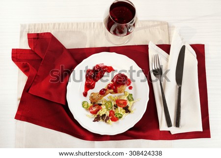 Dish of baked chicken leg and vegetable salad in white plate with glass of wine on table with napkin, top view