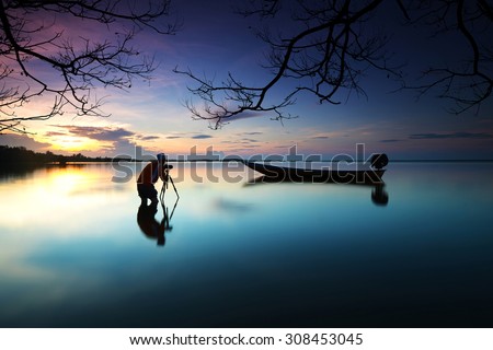 Silhouette of photographer looking to the boat near the beach when the sun goes down with branches tree border. Nature Photography Royalty-Free Stock Photo #308453045