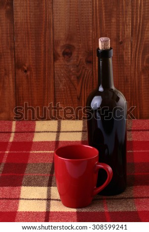 One glass bottle and red cup full of white hot mulled wine standing close to each other on chequered plaid on brown wooden wall background, vertical picture