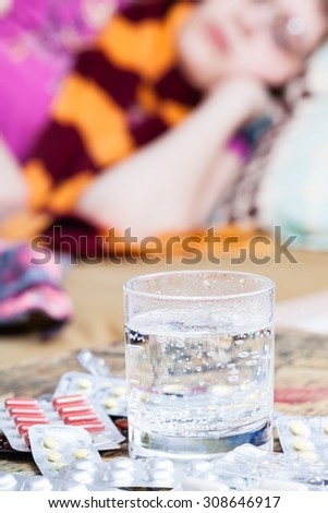 glass with medicament and pile of pills on table close up and sick woman with scarf around her neck on sofa in bed-sitting room on background