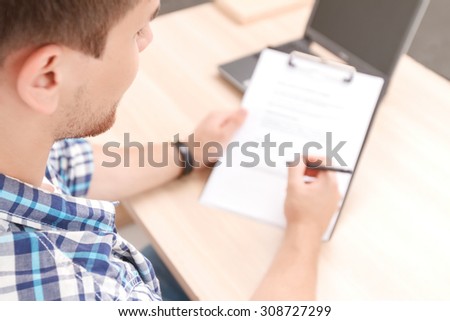 Top view of a young student sitting at the table holding a folder and filling some information for his project work 