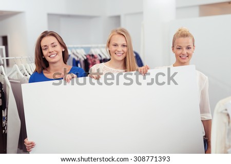 Three Pretty Young Woman Holding Large Clean Poster with Copy Space inside the Clothing Store and Smiling at the Camera.