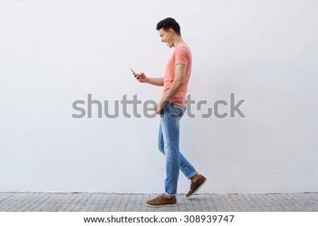 Full length side portrait of a cheerful young man walking on sidewalk and reading text message on mobile phone Royalty-Free Stock Photo #308939747