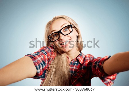 Close up portrait of a young joyful blonde girl holding a smartphone digital camera with her hands and taking a selfie self portrait of herself standing against blue background