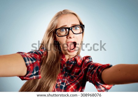 Close up portrait of a young shocked blonde girl holding a smartphone digital camera with her hands and taking a selfie self portrait of herself standing against blue background