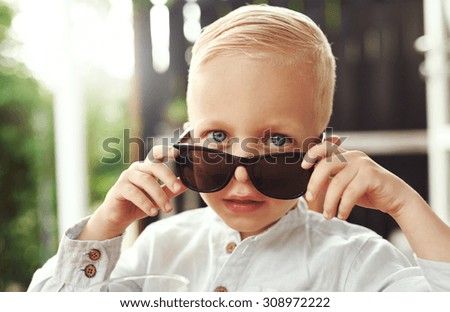 Handsome little boy in trendy over sized sunglasses borrowed from his father peering over the top with penetrating serious blue eyes at the camera