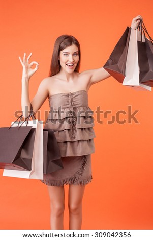 Beautiful girl is going shopping with pleasure. She is showing packets of bought things. The lady is showing okay sign and smiling. Isolated on orange background