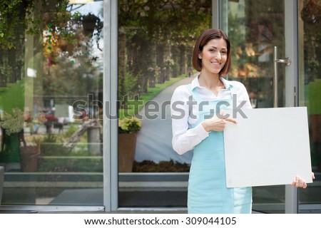 Cheerful saleswoman is standing near her flower shop. She is holding an empty white billboard and pointing her finger at it. The woman is looking forward and smiling. Copy space in left side