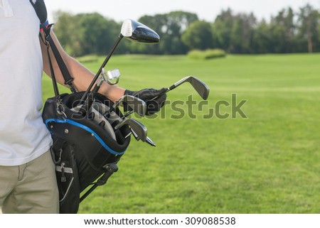Close-up picture of golf bag hold by a golfer. Man in white T-shirt and cream trousers taking a driver to start his game over golf course.