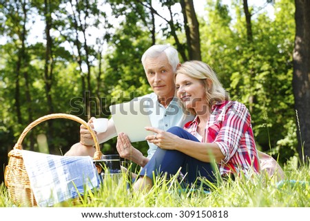 Portrait of senior couple having picnic in the forest. Elderly woman holding hand digital tablet and sitting next to her old man and reading the map online together on the tablet. 