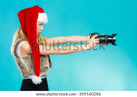 Photographer girl shooting images. Attractive blonde woman wearing santa claus helper hat taking photos with camera. Vivid blue background