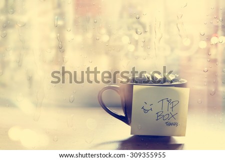 Tip box, coin in the coffee cup in cafe front of mirror and rain water drop, Vintage color and soft concept