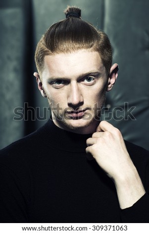Portrait of handsome stylish young unshaven boy with red hair with ponytail standing in black jersey with hand near neck looking forward indoor on studio background, vertical picture