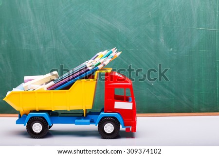 Colorful pencils of red yellow orange violet purple pink green blue chalk and fan english alphabet in plastic truck car toy standing on white school desk on blackboard backdrop, horizontal photo