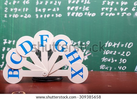 Stationary cup and english alphabet fan with blue capital letters standing on old brown school desk on written with white chalk multiplication table on math lesson blackboard, horizontal picture