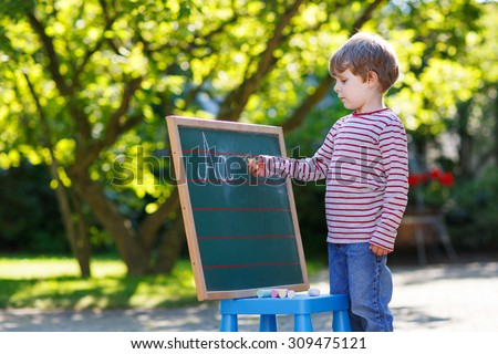 Little boy at blackboard practicing writing letters, outdoor school or nursery. Back to school concept. Lifestyle, kids, education.