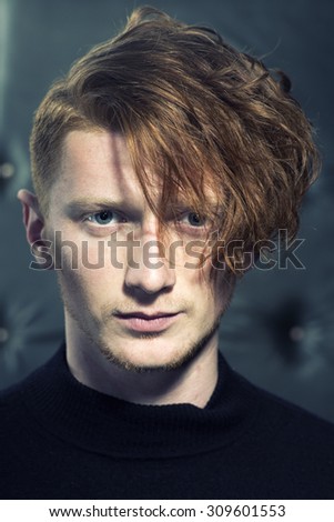 Portrait of attractive stylish young unshaven boy model with red hair standing in black jersey looking away indoor on studio background closeup, vertical picture
