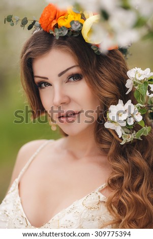 Bride in blooming apple trees. Woman in flower wreath and wedding bouquet in hands. Beautiful bride in wedding dress posing in a blooming apple garden. Spring mood. Young woman outdoors
