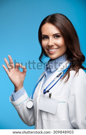 Portrait of happy smiling young female doctor showing okay hand sign, on blue background