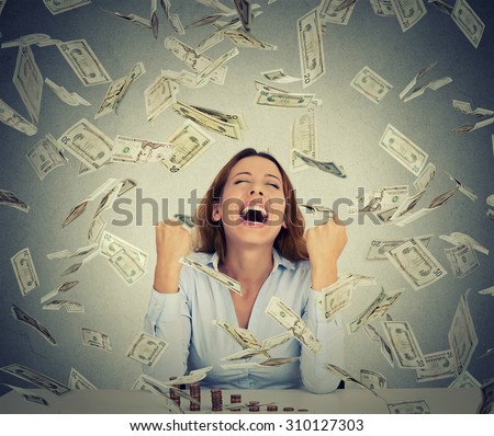Excited happy young woman sitting at table with growing stack of coins under a money rain isolated on gray wall background. Positive emotions financial success luck good economy concept