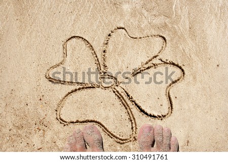 Clover symbol drawn in sand on a beach in an exotic island background with feet  for summer, ocean, sea, travel, vacation, tourism, tropical, coast, message, resort, paradise, sunny or water