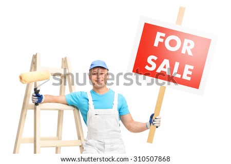 Young male painter posing next to a wooden ladder and holding a paint roller in one hand and a for sale sign in the other isolated on white background