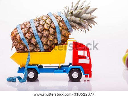 Plastic colorful toy truck yellow blue and red colors with one fresh big pineapple fruit and measuring tape as diet symbol in basket on white studio background closeup, horizontal picture