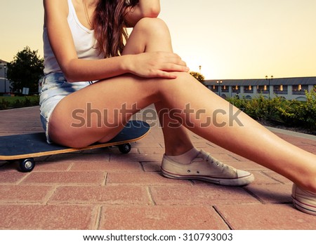 Legs of skateboarder girl sit on the board in short jeans shorts place for text is on the ground. Toned color image. Colorized. Long legs of the women.