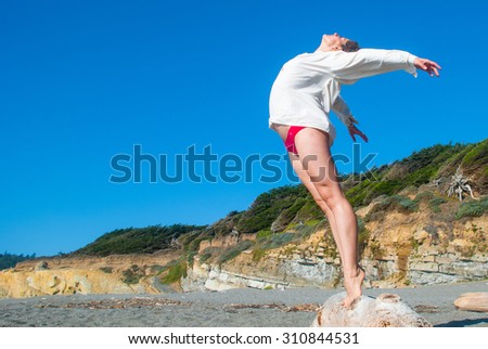 A young woman with strong long legs and wearing a flowing white blouse and red panties tiptoes on the edge of a driftwood log and thrusts her torso into the breeze on a beach in northern California.  