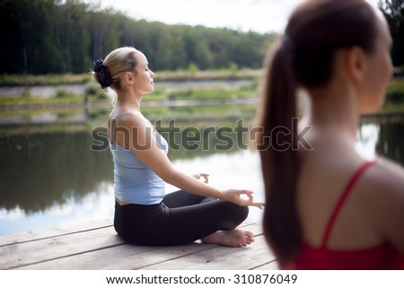 Two serene beautiful sporty fit young women working out on the lake on summer day, Sitting in Easy (Pleasant Posture), Sukhasana, meditating with closed eyes, breathing, back view Royalty-Free Stock Photo #310876049