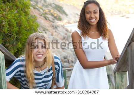 Beauty portrait of two young teenager girls of different ethnic origins friends smiling together by the sea on a summer holiday, outdoors. Travel fun and adolescent lifestyle, nature beach exterior.