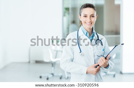 Smiling female doctor with lab coat in her office holding a clipboard with medical records, she is looking at camera Royalty-Free Stock Photo #310920596