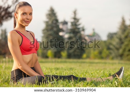 Beautiful sportswoman is sitting on grass and doing exercises. She is smiling and looking at the camera with joy.  Copy space in right side