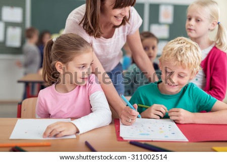 Smiling female teacher helping a young boy at primary school with his notes watched by two young girls Royalty-Free Stock Photo #311002514