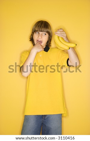 Portrait of Caucasian boy holding bunch of bananas and gesturing with finger in mouth that they are gross standing against yellow background.