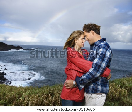 Caucasian mid-adult couple embracing by ocean with rainbow in background in Maui, Hawaii.