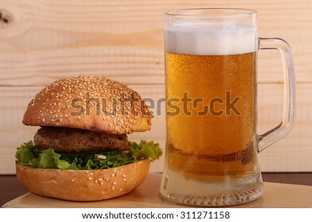 Big fresh tasty burger of green lettuce meat cutlet tomato and white bread bun with sesame seeds near and glass of light beer with froth on octoberfest holiday on wooden background, horizontal picture