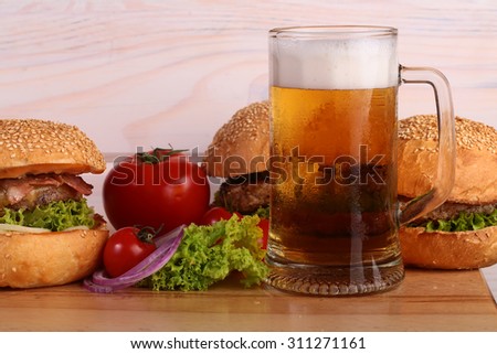 Few fresh tasty burgers of green lettuce meat cutlet tomato and white bun with sesame seeds near and glass of light beer with froth on octoberfest holiday on wooden background, horizontal picture