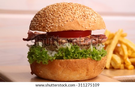 Big tasty appetizing fresh burger of green lettuce red tomato cheese and bacon slice meat cutlet and white bread bun with sesame seeds and chips closeup, horizontal picture