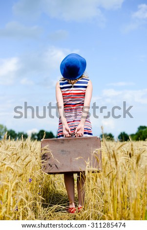 Picture of lady with navy blue hat holding suitcase in rural countryside, looking away and thinking where to go on sunny blue sky background