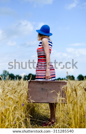Picture of female with suitcase in full length in rural countryside. Lady with navy blue hat on looking away and thinking where to go on sunny day background